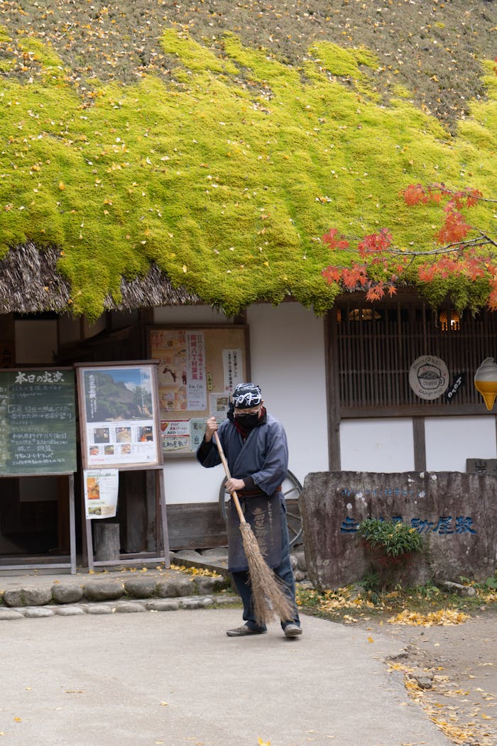 A gardener with a broom in traditional attire cleaning outside a thatched-roof house in Toyota, Japan.